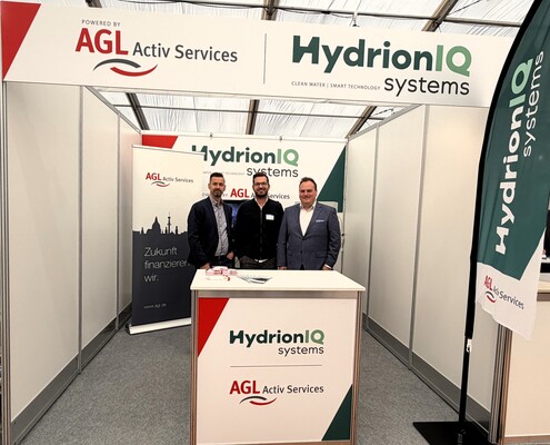 AGL und HydrionIQ auf der Wassertagung von Mösslein Wassertechnik Messestand von AGL Activ Services und HydrionIQ Systems auf der Wassertagung von Mösslein Wassertechnik. Drei Unternehmensvertreter stehen hinter dem gemeinsam gebrandeten Stand. Sichtbar sind die Logos beider Unternehmen auf Bannerflächen, Roll-ups und einer Beachflag. Der Stand präsentiert die Partnerschaft zwischen innovativer Wasseraufbereitung und passenden Finanzierungslösungen für die Wasserwirtschaft.